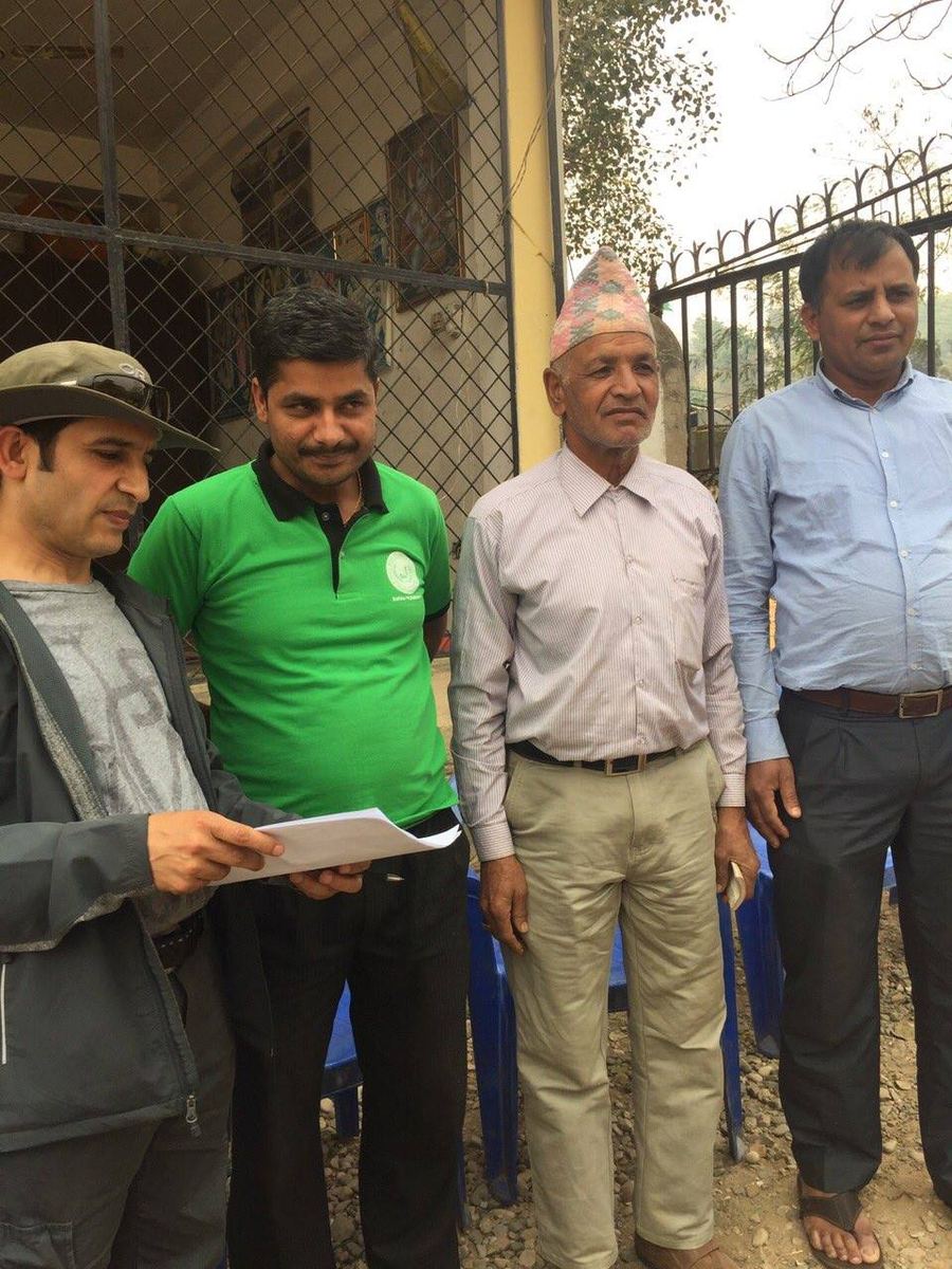 Four men stand together outside a building, one in a green shirt holding papers and reading from them.