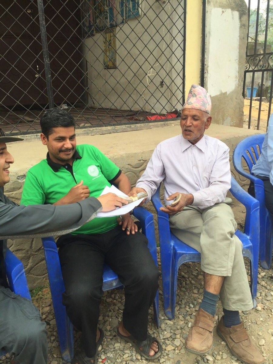 A man in a green shirt hands papers to an elderly man in a Dhaka topi seated on a blue plastic chair in a yard.