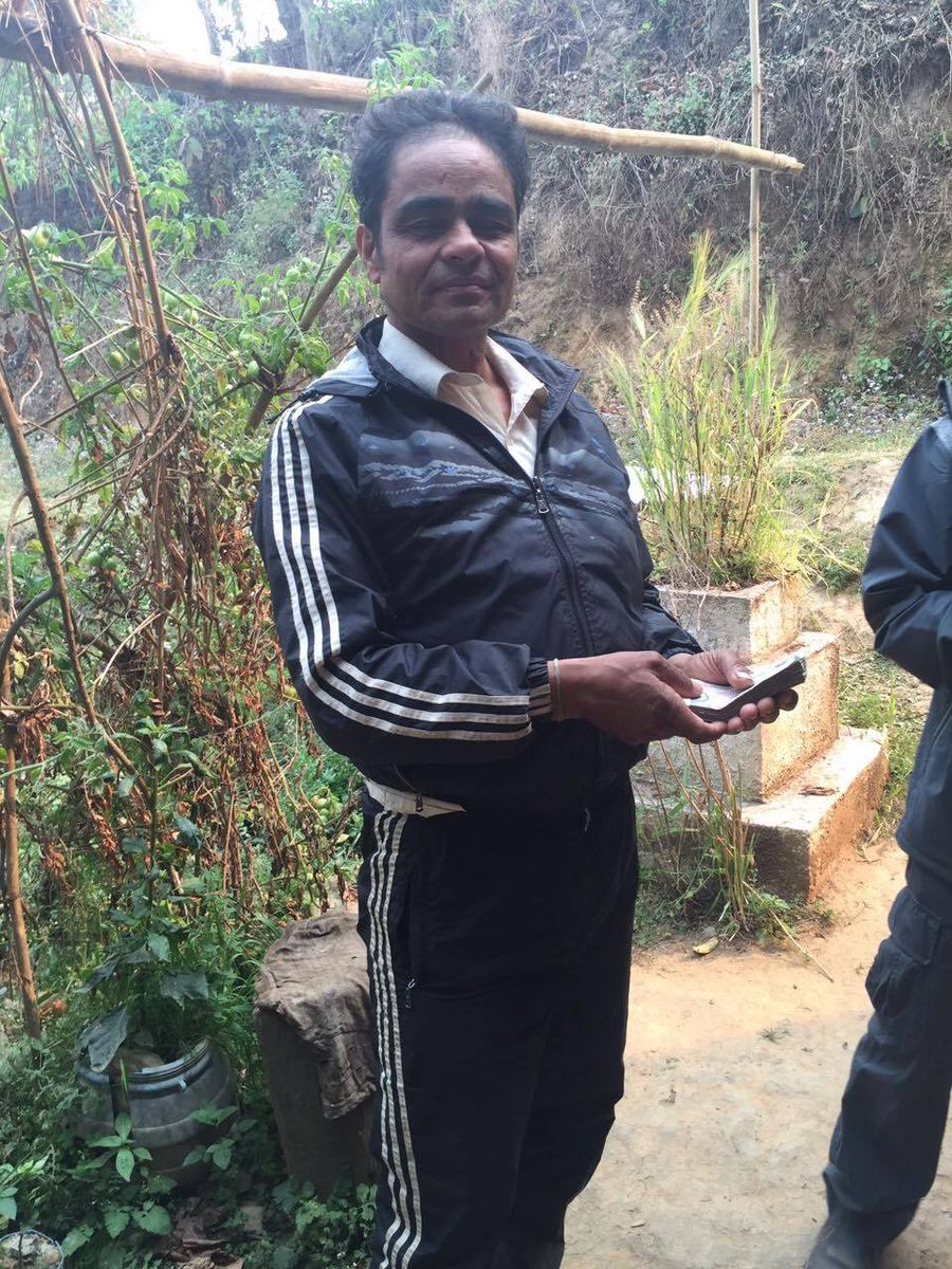 A man in a black tracksuit stands on a hill path holding a phone, with scrub and a stone wall behind him.