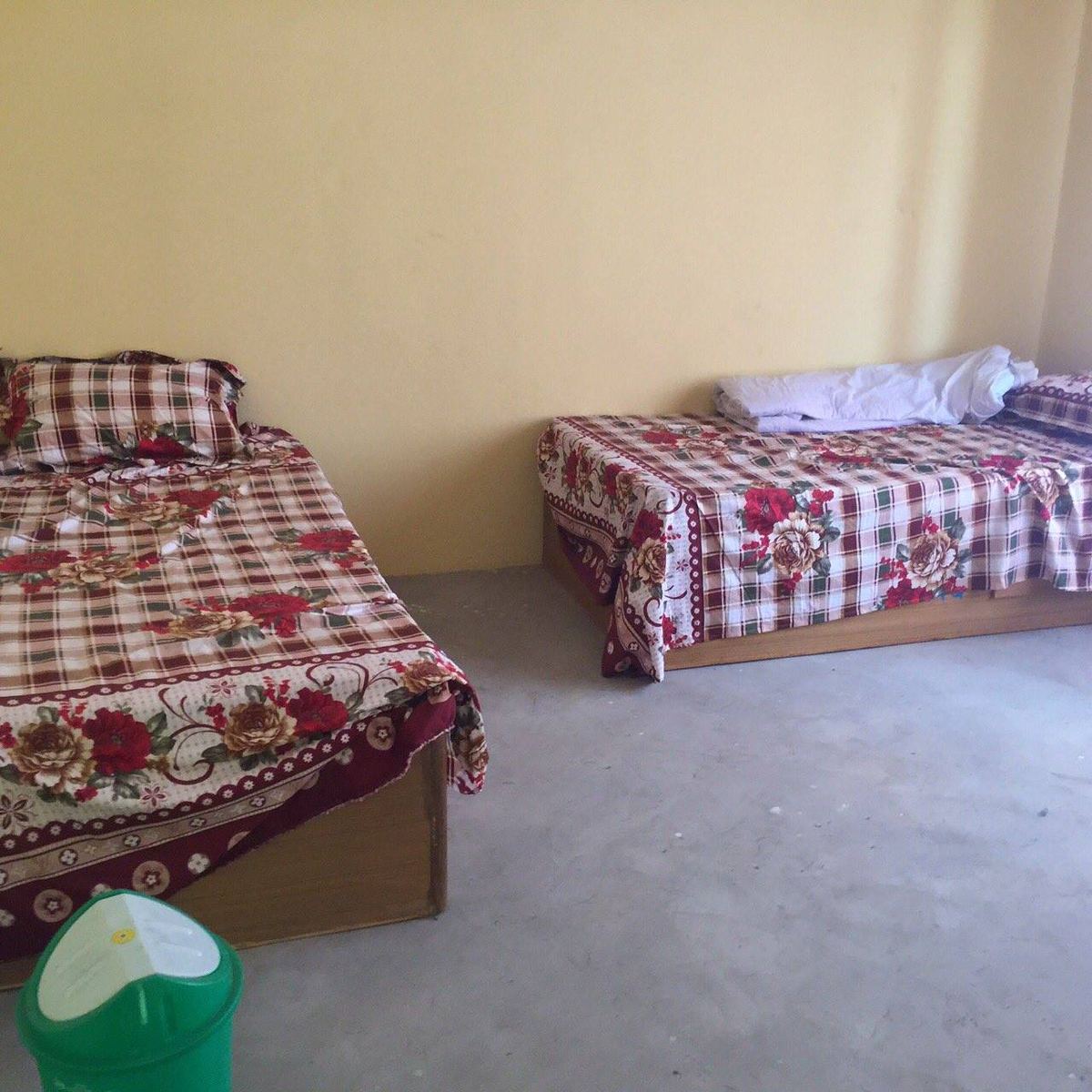 A bare room with a concrete floor and two beds made up with floral covers, and a green water container on the floor.