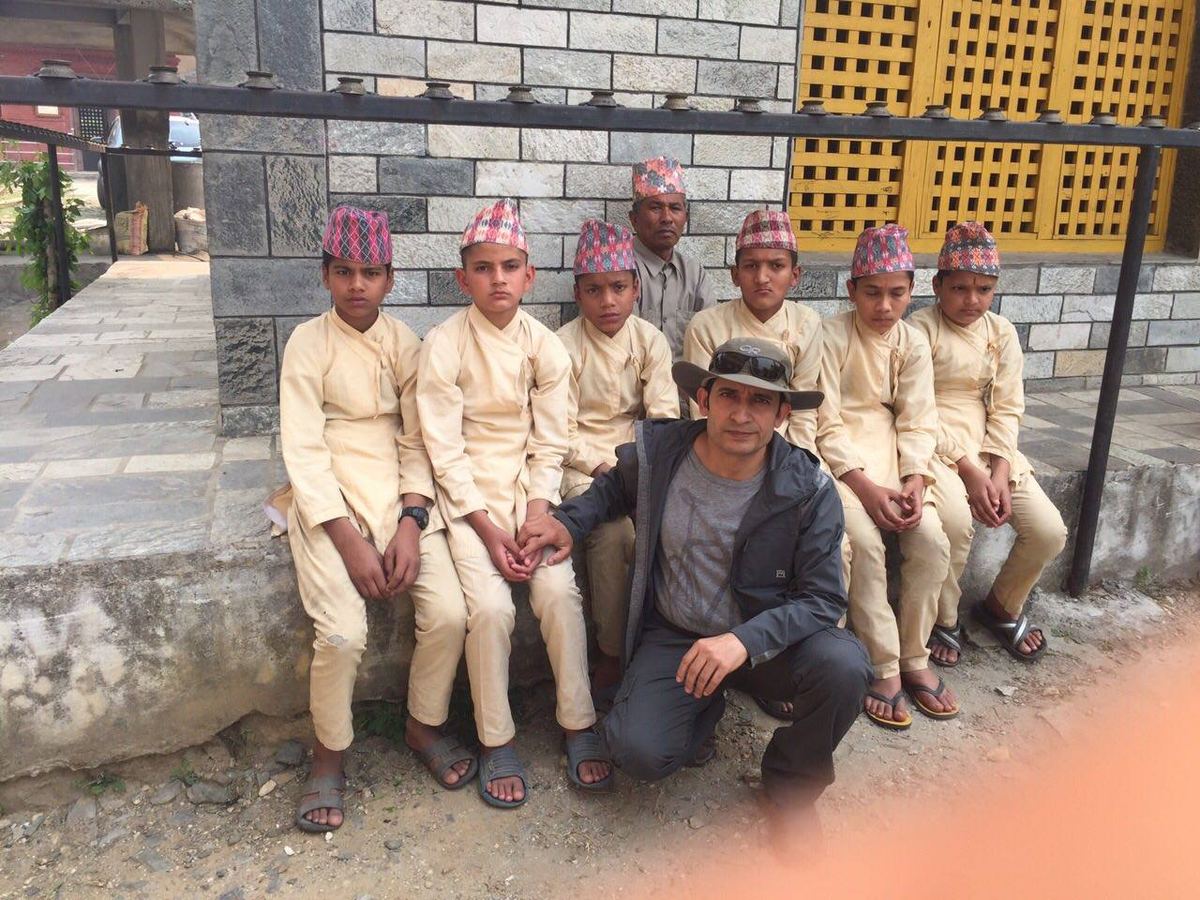 Eight boys in cream kurta and Dhaka topi stand along a low wall beside a new stone and block building, with a man crouching at the end of the row.