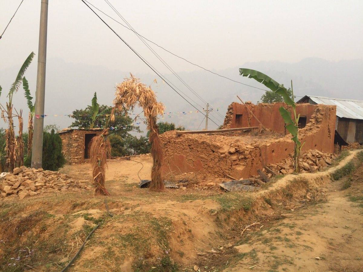 The ruined walls of a mud house on a bare hillside, roof gone, with power lines overhead and hills in the haze behind.