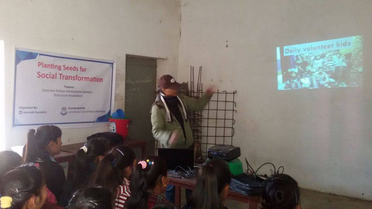 A speaker in a cap and green jacket stands beside a projected slide in a bare classroom, addressing schoolchildren seated on the floor.