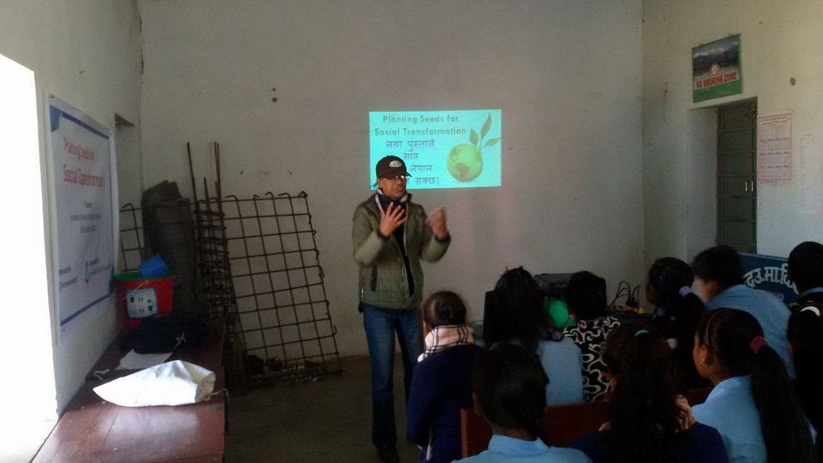 The speaker gestures towards a small printed poster taped to a classroom wall while students watch from the floor.