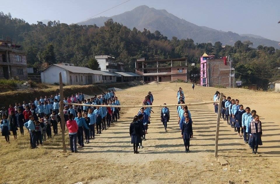 Several hundred pupils in blue uniforms stand in long lines across a bare school ground, with a forested mountain ridge behind.