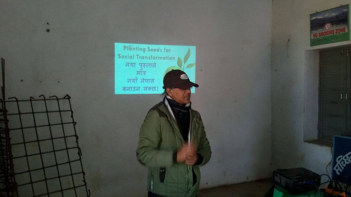 The speaker stands at the front of a classroom beside a poster, one hand raised, with a projector and equipment on a table beside him.