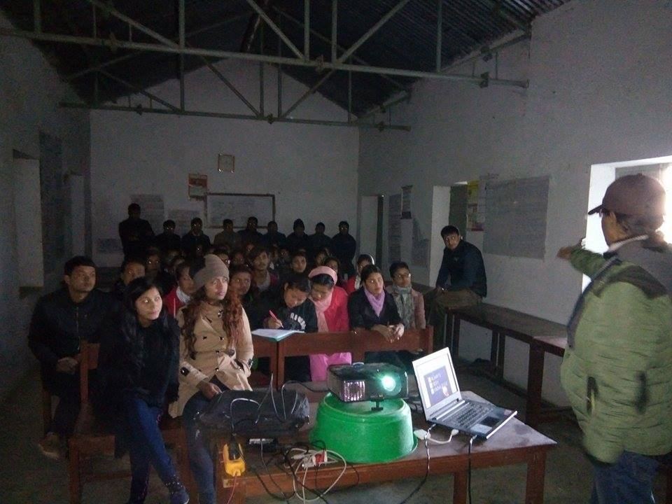 Students sit on benches in a dim hall watching a projector set up on a table, with the speaker standing at the side.