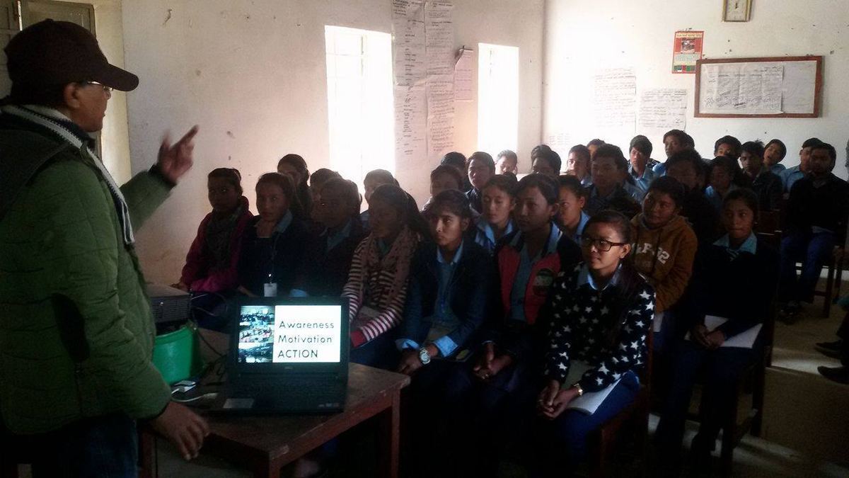 The speaker points across a full classroom of students seated at desks, many holding papers, with a laptop open in front of him.