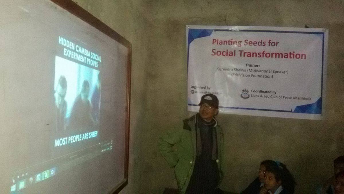 A darkened room with two projected images on the wall, one a slide reading Planting Seeds for Social Transformation, the speaker standing between them.