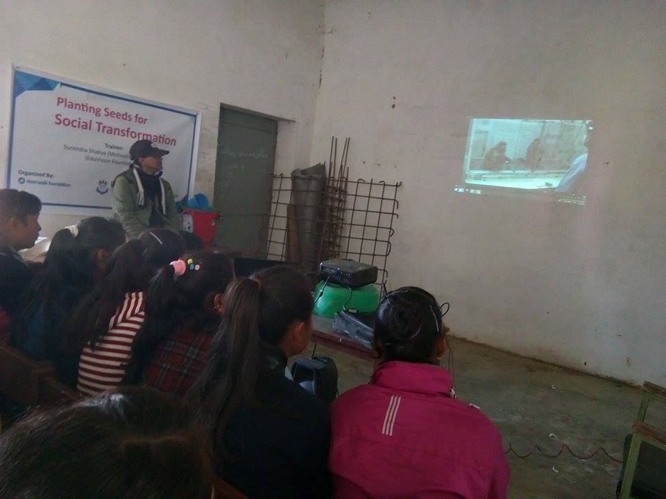Students seated on a classroom floor watch a projected video, the session poster taped to the wall beside the screen.