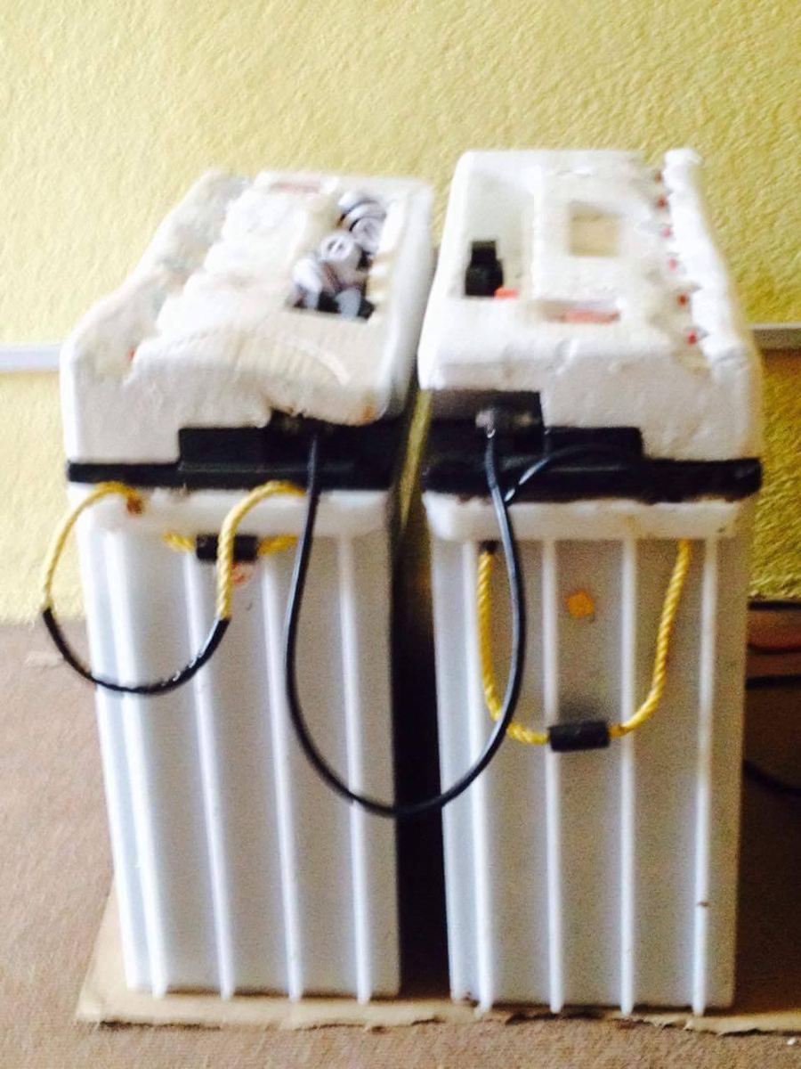 Two tall white lead acid batteries in polystyrene packing on a floor, their terminals wired together.