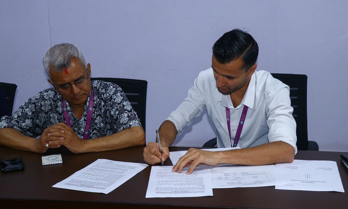 Two signatories seated at a table, one signing the memorandum of understanding while the other reads a copy of the document.