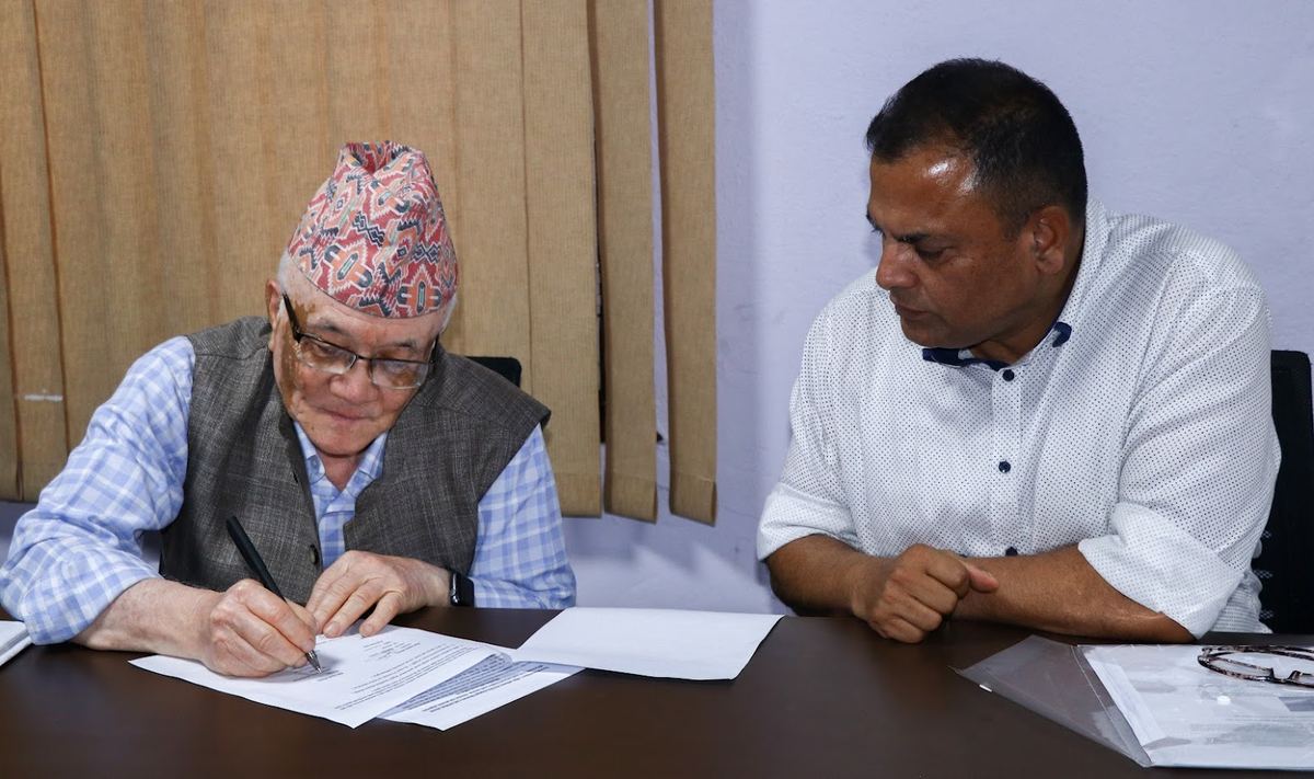 A signatory wearing a Dhaka topi signs the memorandum at the table while a colleague seated beside him looks on.