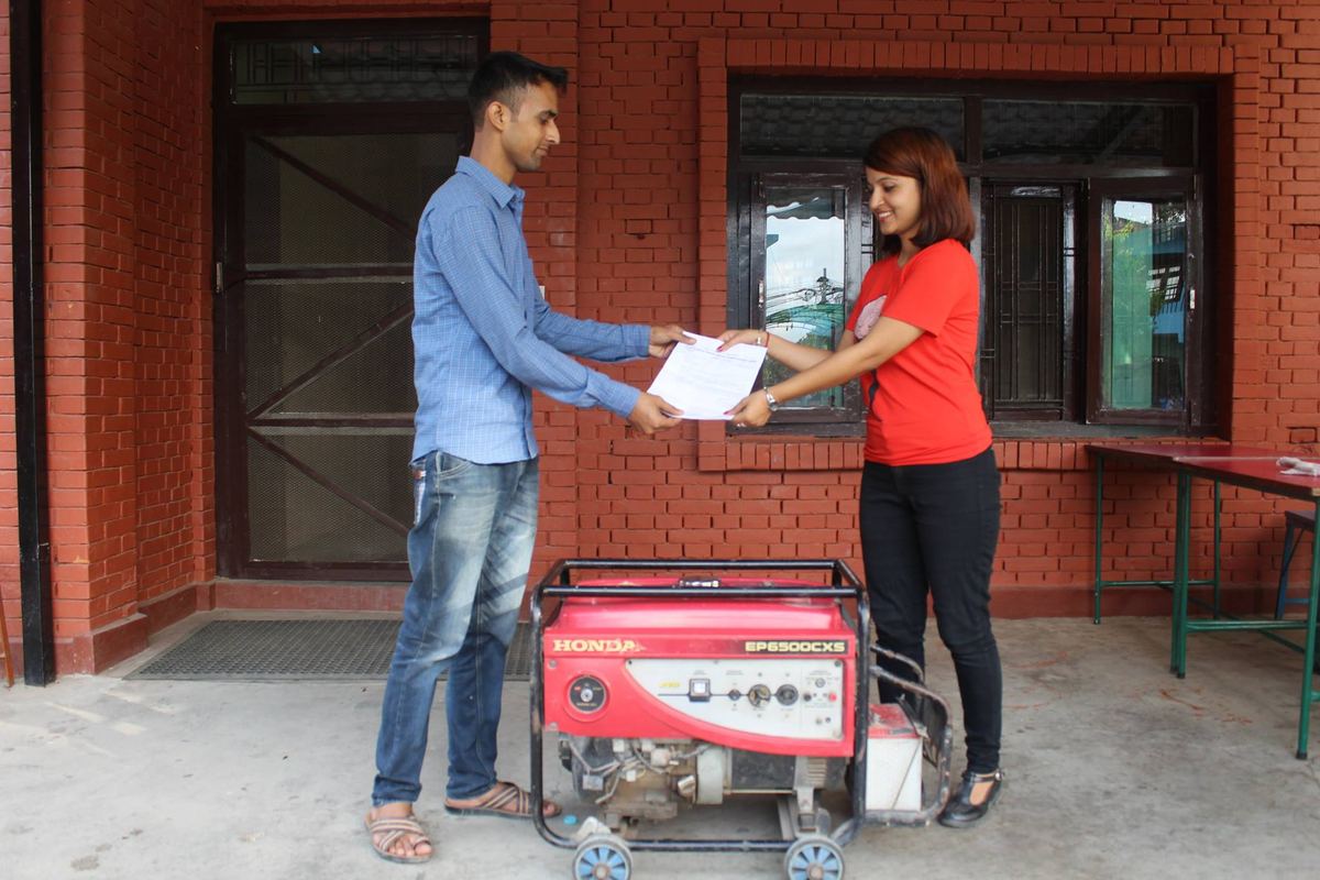 A man in a blue shirt and a woman in a red t-shirt hold a document between them over a red Honda generator, outside a brick building.