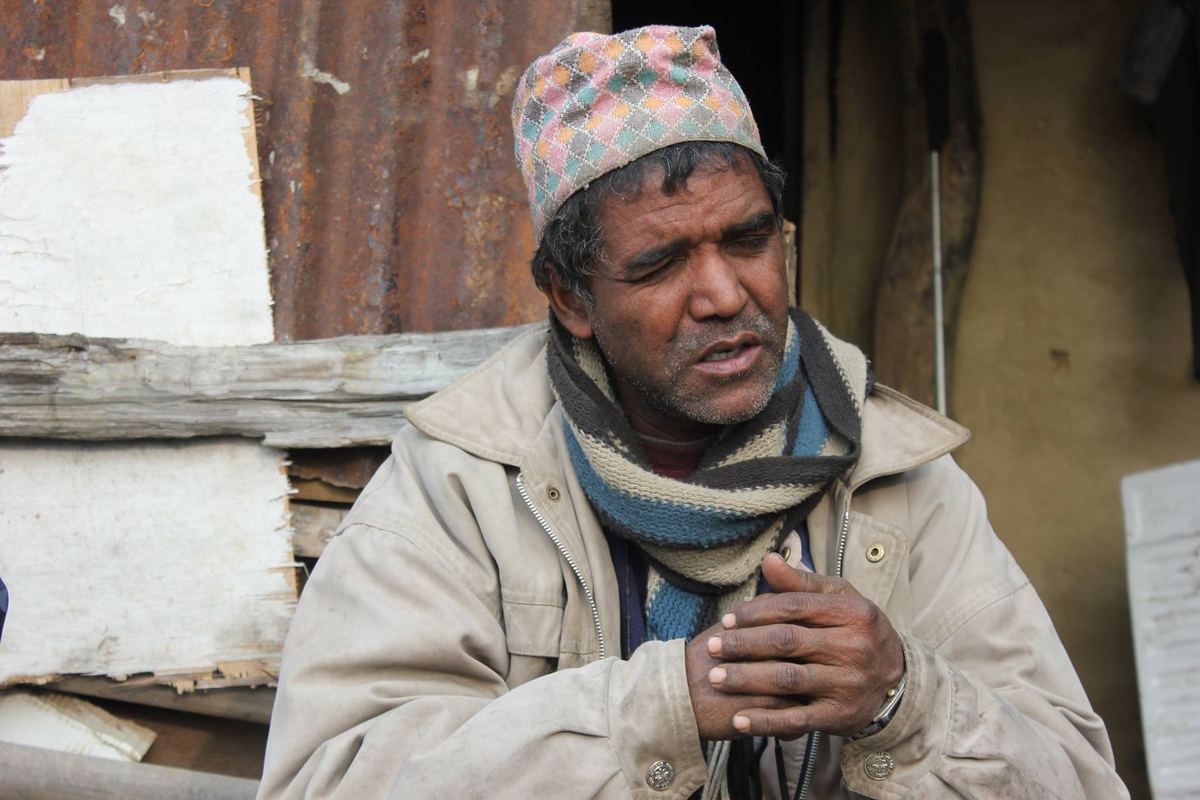 Portrait of a man in a Dhaka topi and cream coat seated outside a mud walled house, hands clasped in front of him.