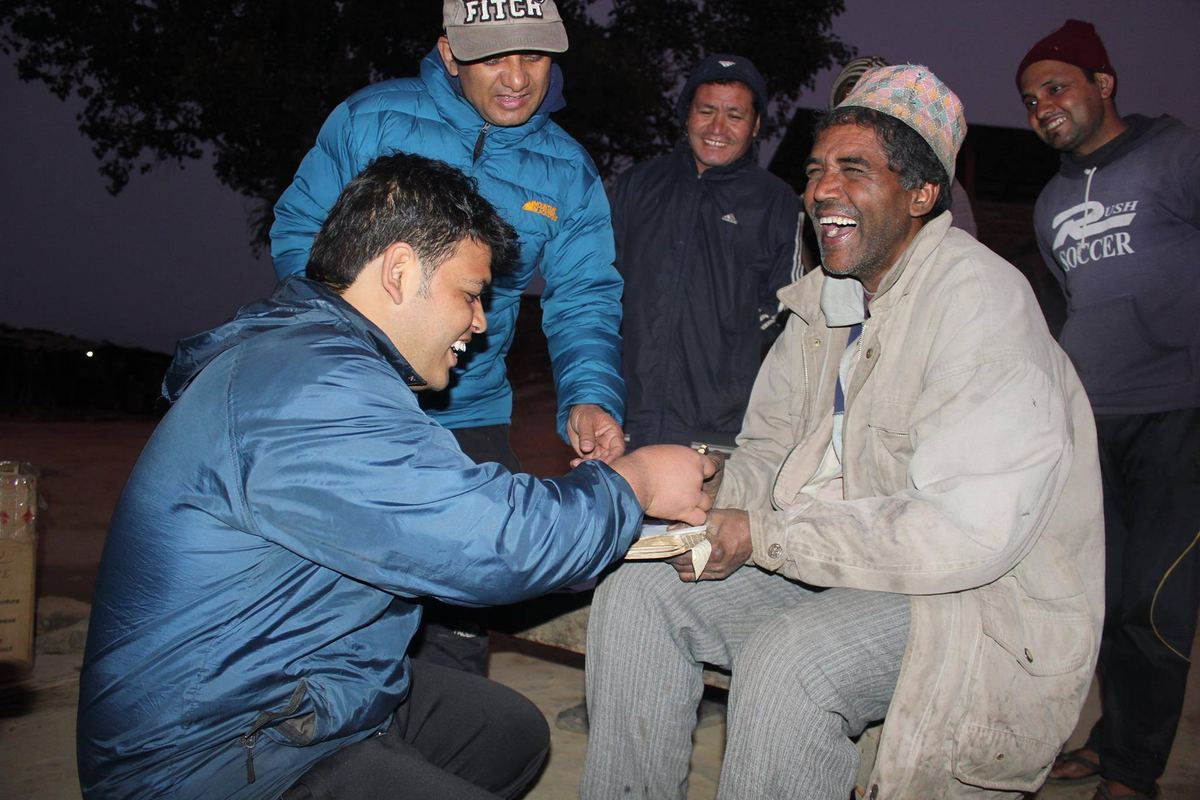 A man in a blue down jacket crouches to take the hand of a man in a cream coat and Dhaka topi seated on a bench at dusk, others standing around them.