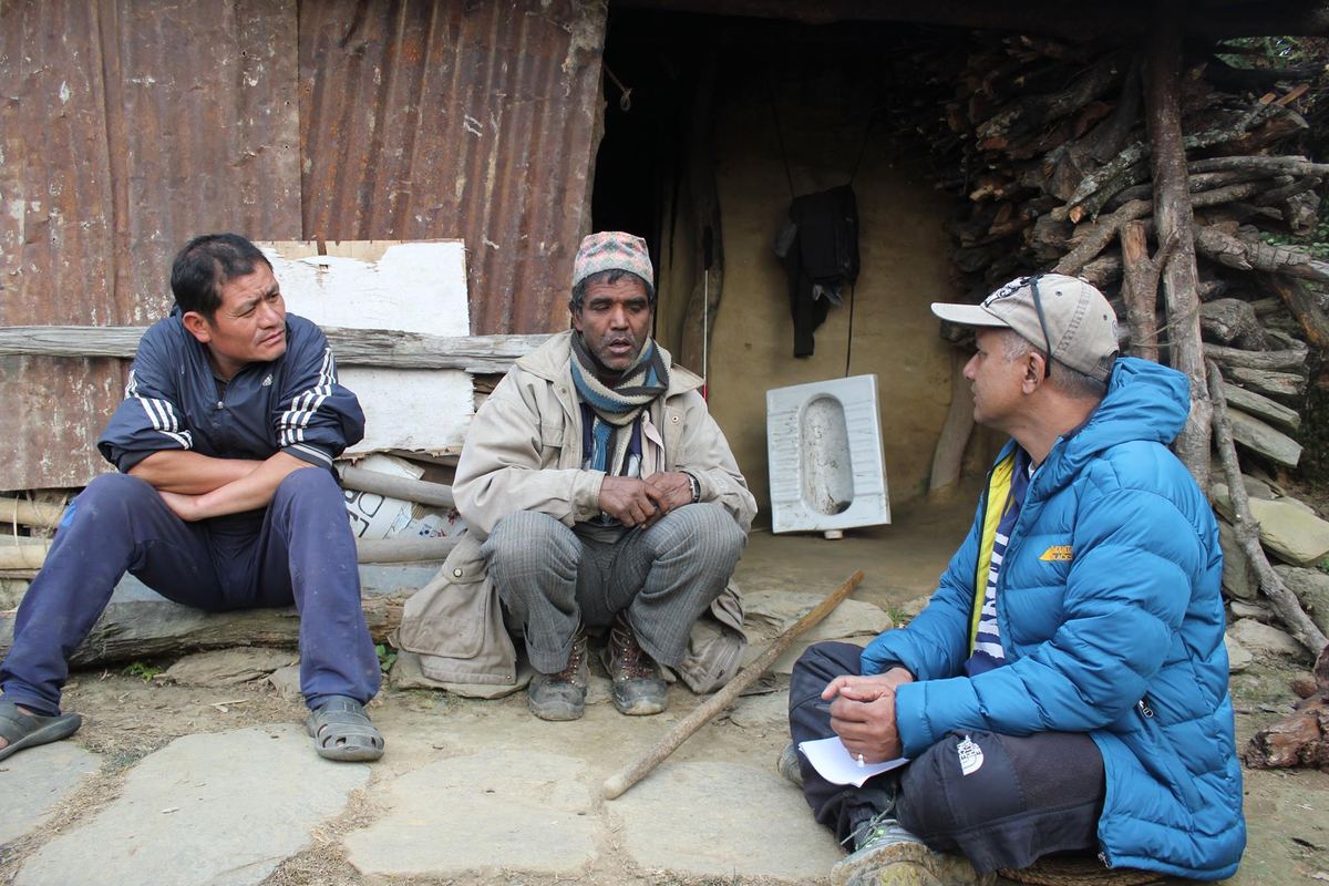 Three men sit talking on a stone step outside a timber house, one in a blue jacket taking notes.