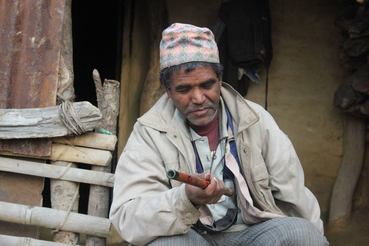 The man sits holding the flute across his lap, one hand open, in front of a mud and timber wall.