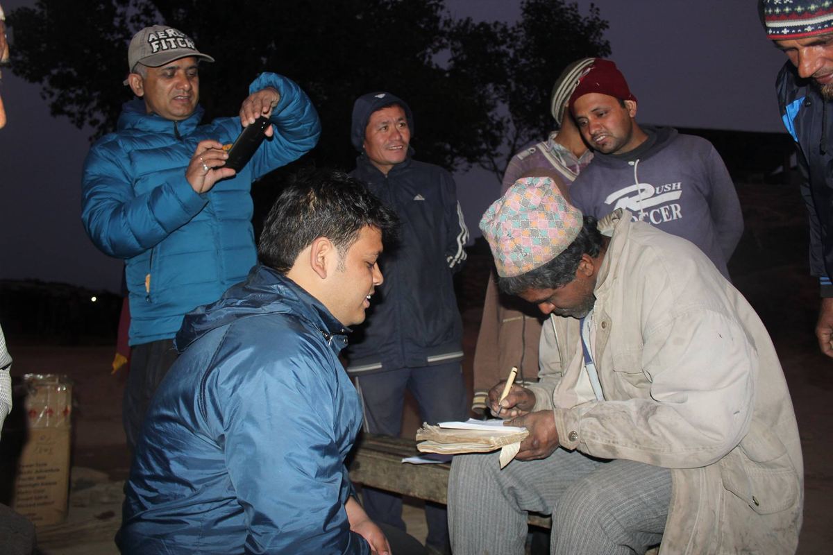 Two men crouch beside the seated porter at dusk, one writing on a pad resting on a bench, villagers watching behind.