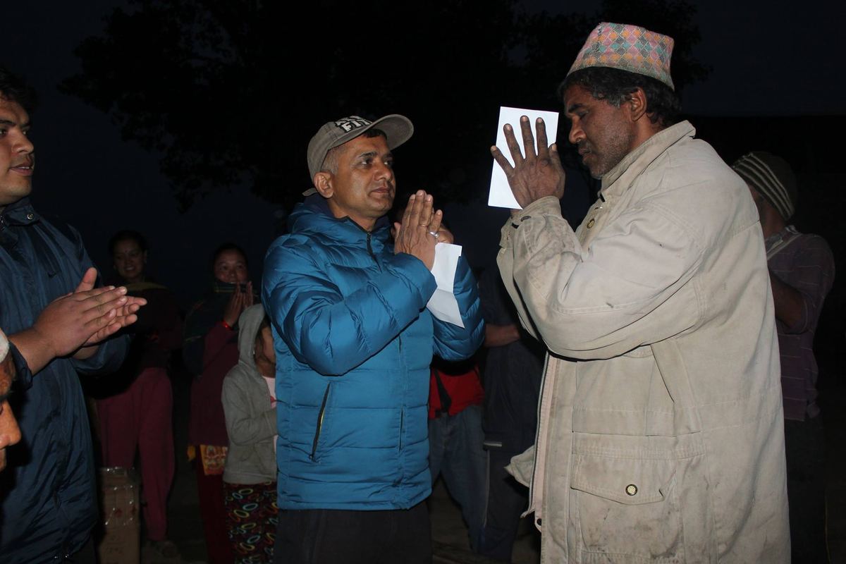 The porter stands with his hands folded in namaste facing a man in a blue jacket who returns the greeting, villagers gathered around.