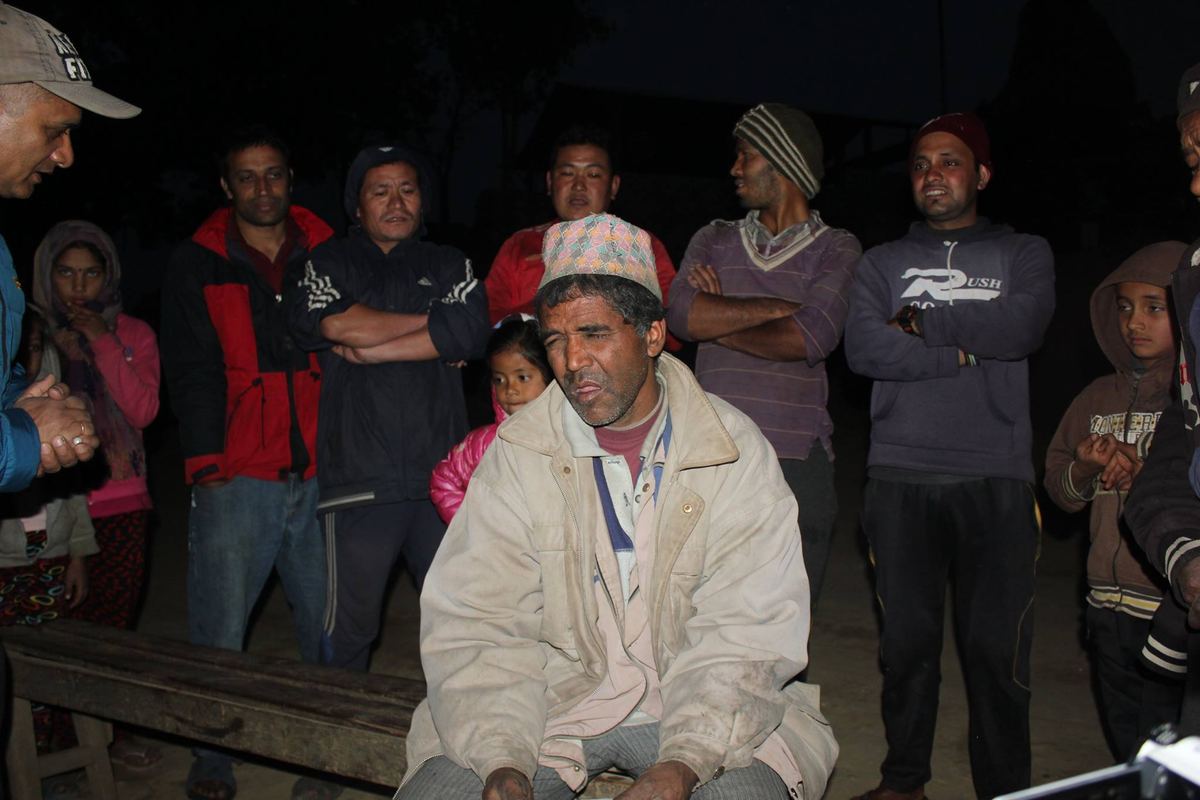 The porter sits on a bench in the dark surrounded by a dozen standing villagers, all looking towards him.