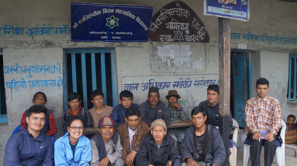 About a dozen people sit and stand in front of a small school building with a Nepali signboard above the door and writing painted on the wall.