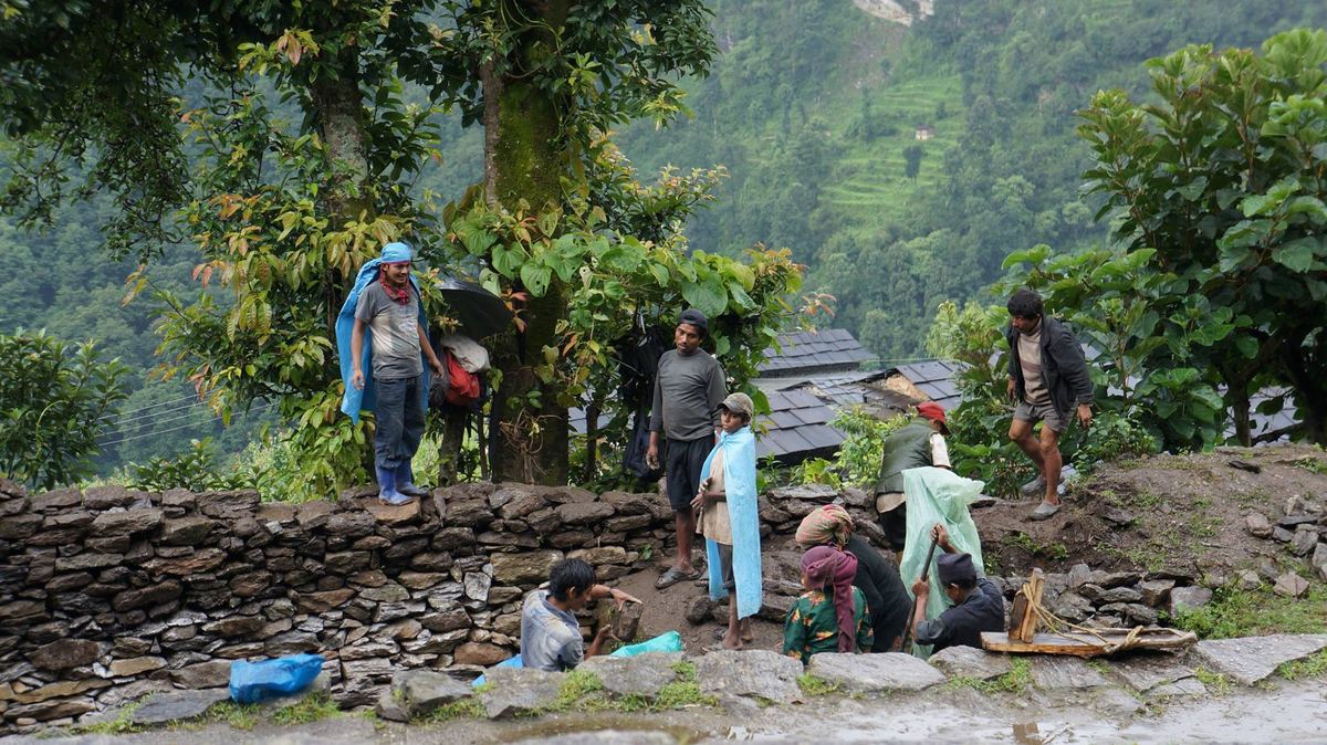 People work on a dry stone retaining wall on a steep green hillside, with bundles and containers set down on the terrace beside them.