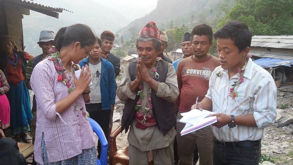 A woman in a pink shawl stands with her hands folded in namaste beside villagers, two men in Dhaka topi returning the greeting and one holding papers, with a river valley behind.