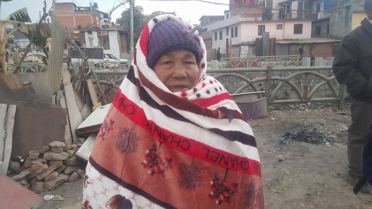 The elderly woman stands wrapped in the new blanket looking at the camera, with a demolished wall and houses behind her.