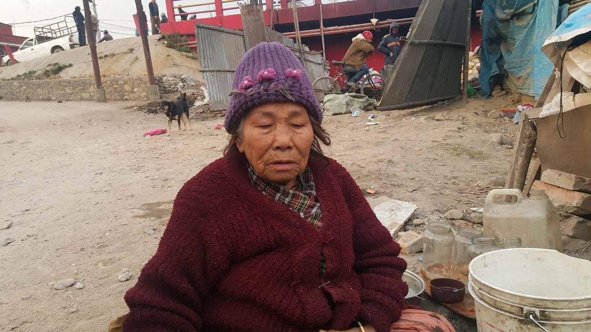 An elderly woman in a maroon jumper and purple woollen hat sits on the ground beside a bucket and a cooking pot amid rubble and scrap.
