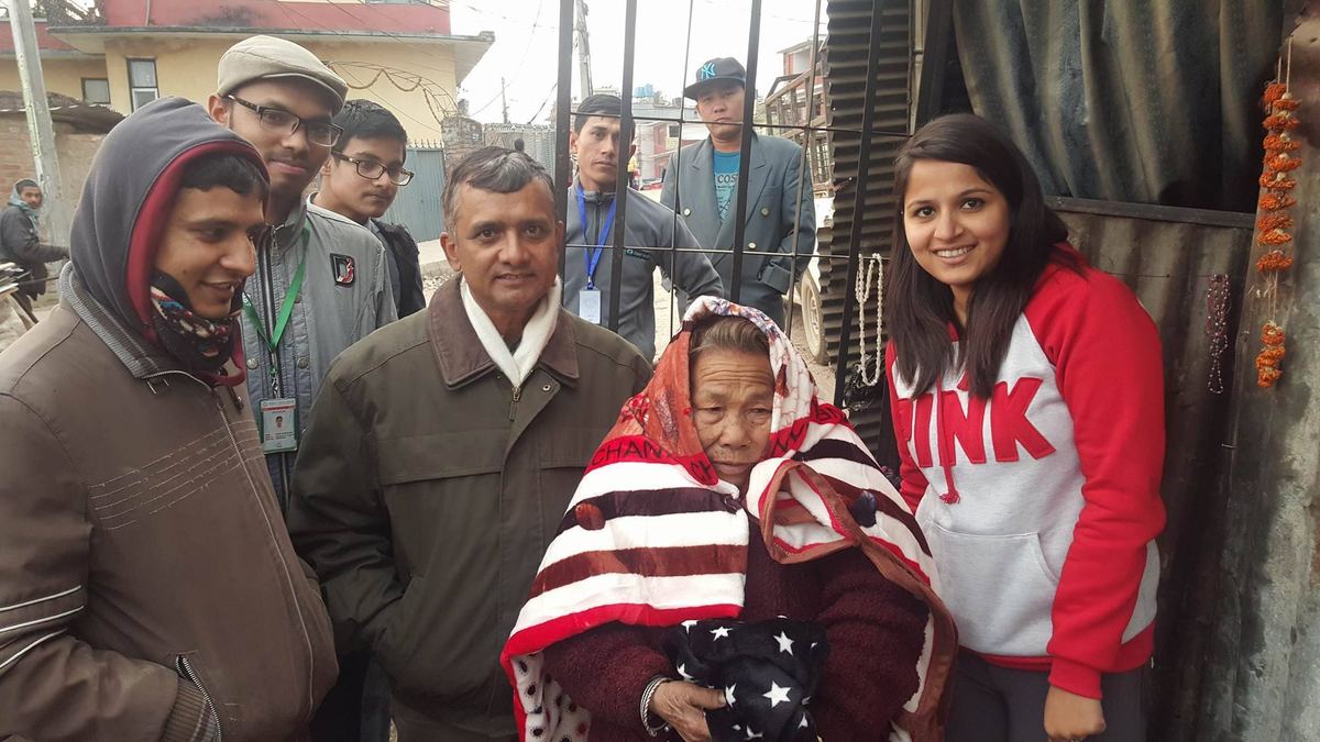 Four visitors stand around the elderly woman outside a corrugated iron shelter, a new patterned blanket draped over her shoulders.