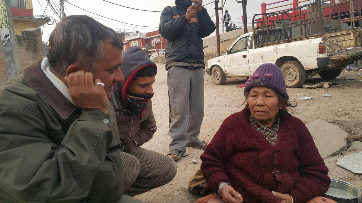 Two men crouch and stand on waste ground talking to the woman, who sits on the ground beside a metal pot, with a pickup truck parked behind.