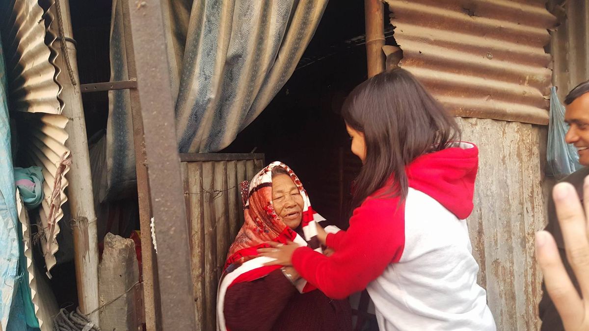 A young woman in a red and white top reaches up to wrap a blanket around the woman standing in the doorway of a tin shelter.