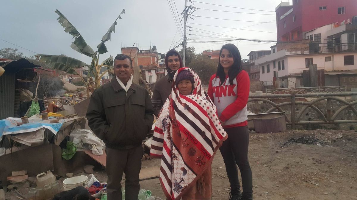 Three visitors stand with the woman on a track between buildings, the blanket around her, with rubble and a banana plant behind.