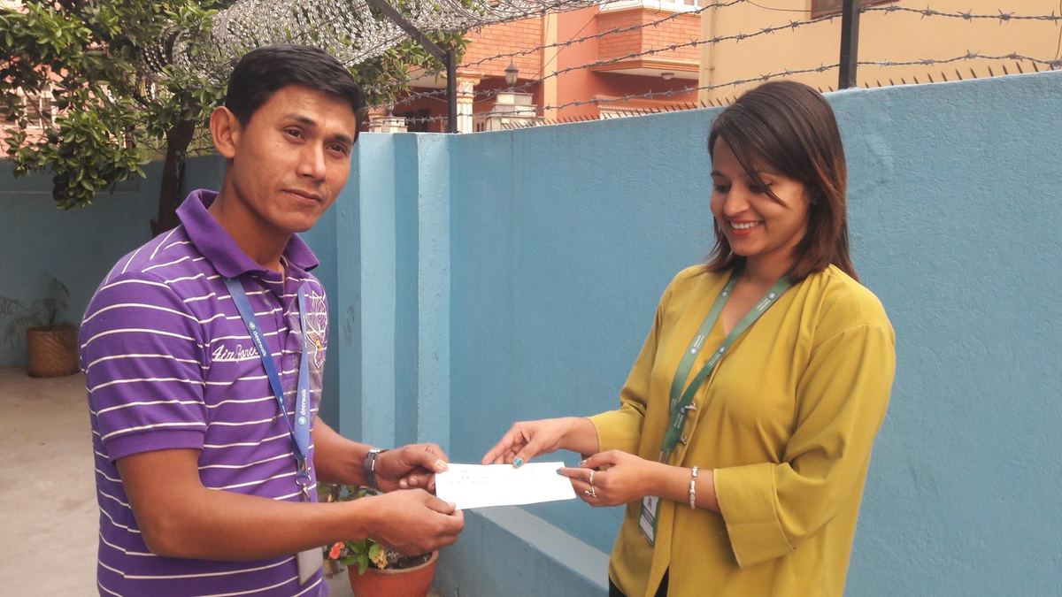 A young man in a purple striped polo shirt hands a white envelope to a woman in a yellow kurta in front of a blue painted wall.