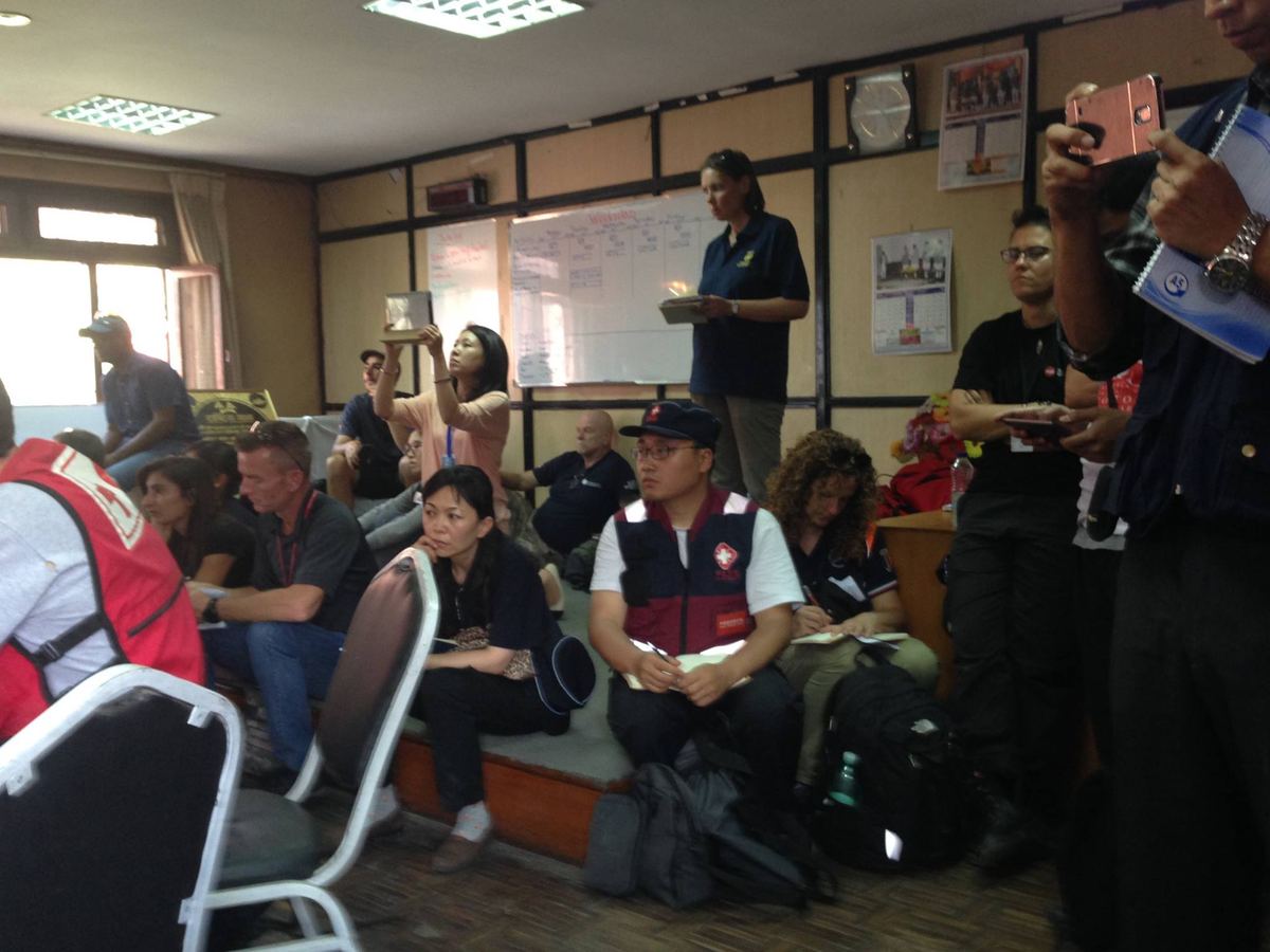 Medical team members in uniform sit and stand around a hall with kit bags at their feet, one addressing the room.