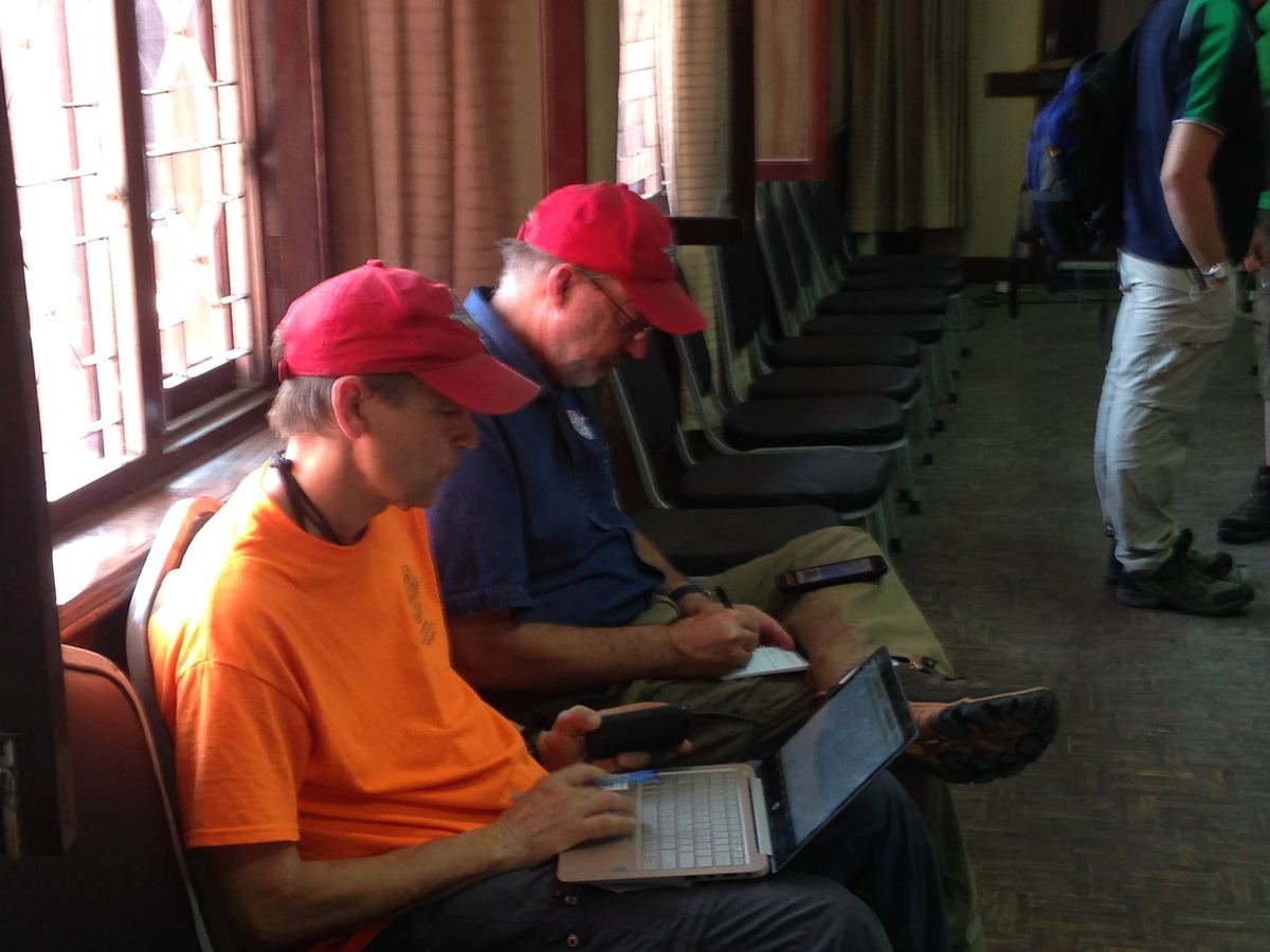 Two men in caps sit in a row of conference chairs working on laptops balanced on their knees.
