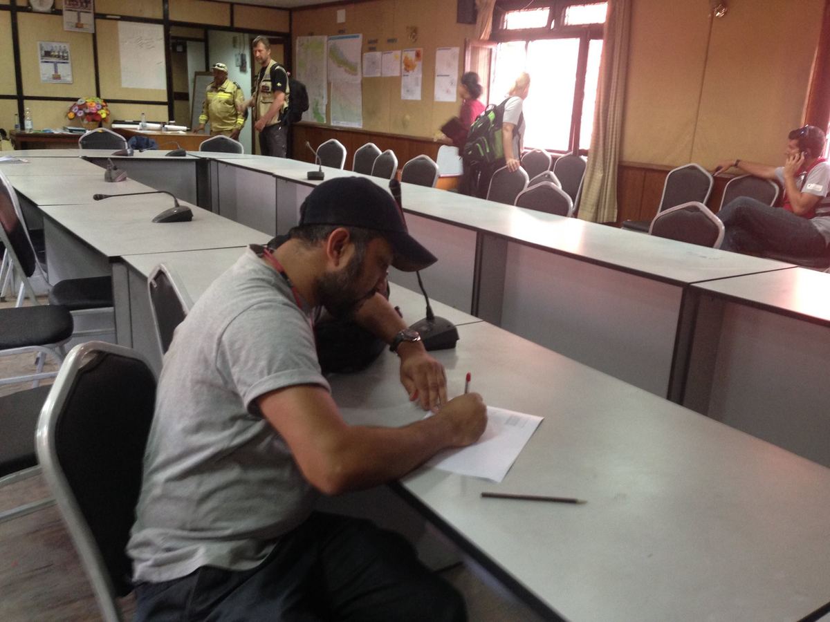 A man in a cap sits alone writing at a long conference table, with the meeting continuing at the front of the room behind him.