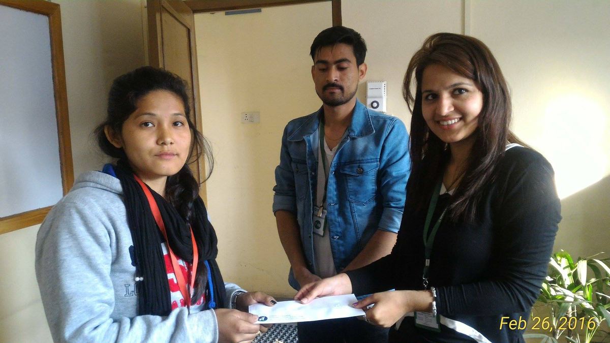 A young woman takes an envelope from a woman in a black jacket in an office doorway, a man standing behind them.