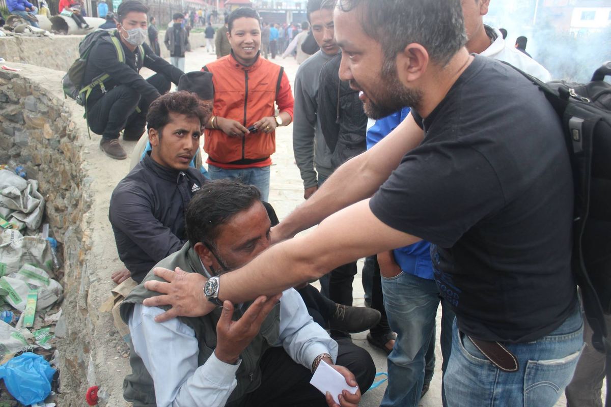 A volunteer leans in to speak to a man seated on a stone wall among a crowd of people, resting a hand on his shoulder.
