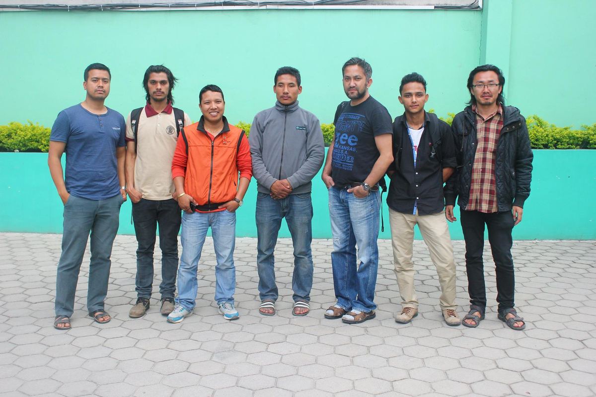 Ten young men stand in a line in front of a turquoise painted wall, several carrying rucksacks.
