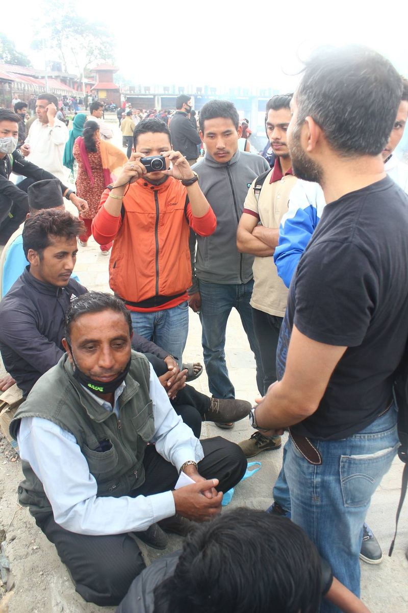 Volunteers crouch and stand talking to people seated on the ground in a crowded open area, one taking a photograph.