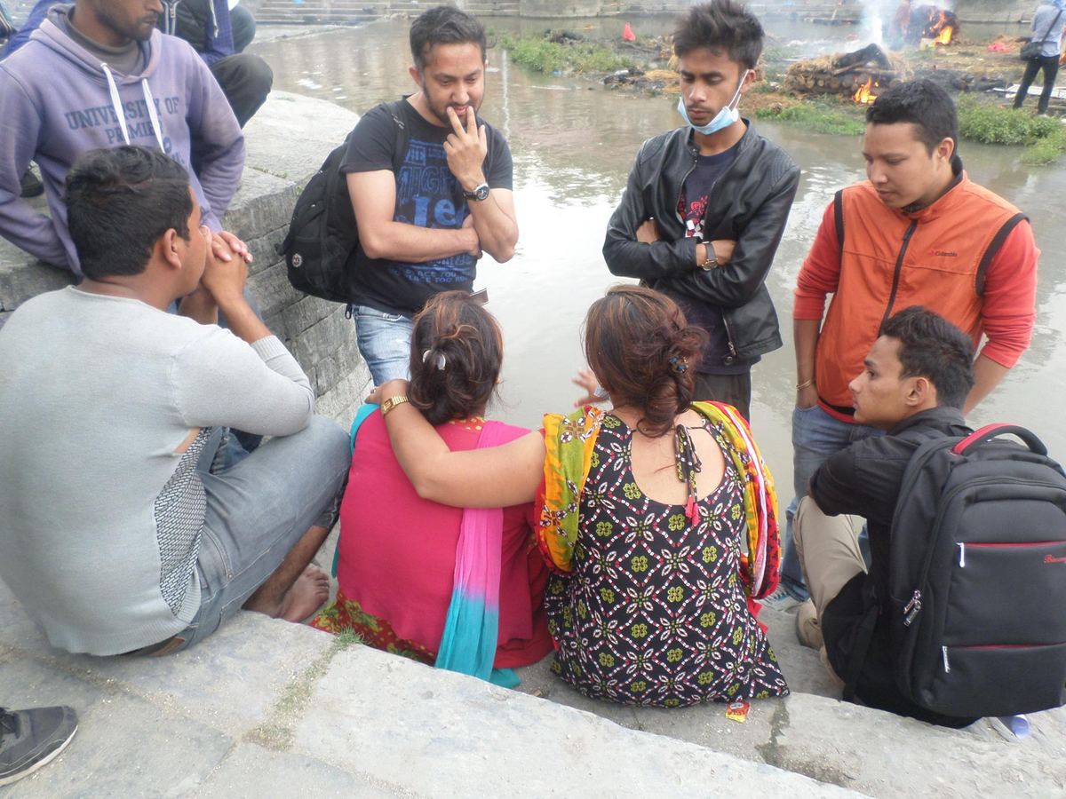 Families sit along a low stone embankment with bags beside them while volunteers stand around them, smoke rising in the distance.