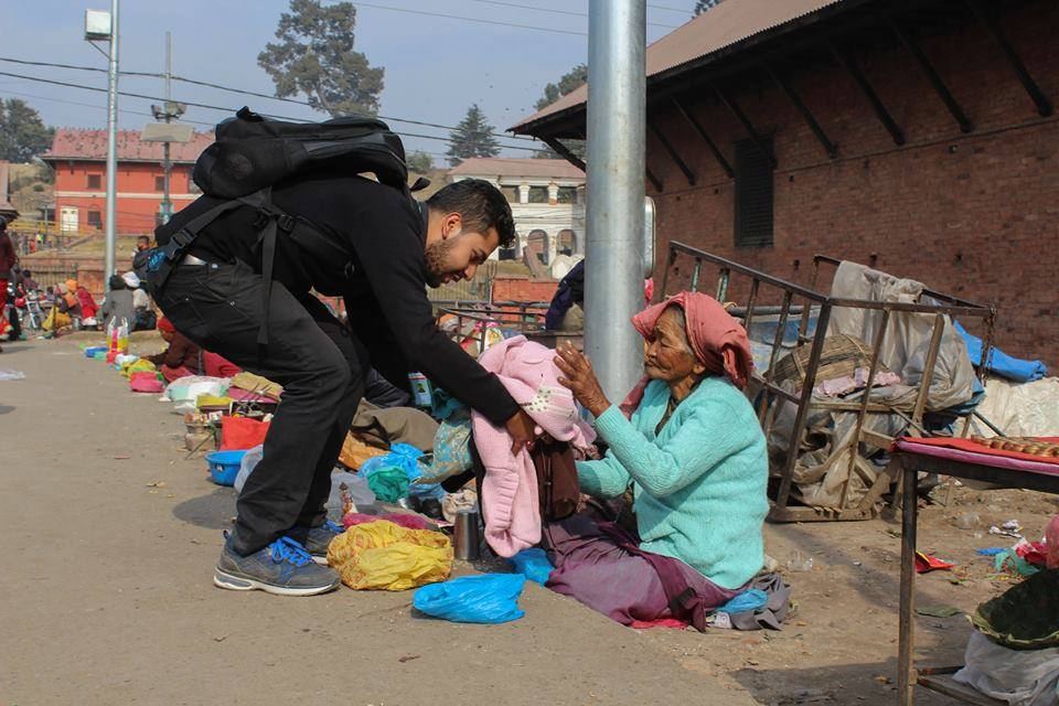 A volunteer crouches to hand clothes to a woman sitting on a street beside her belongings and a handcart.