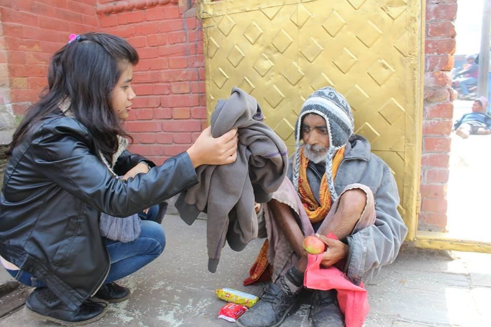 A volunteer holds out a folded grey blanket to two elderly people sitting against a yellow metal gate.
