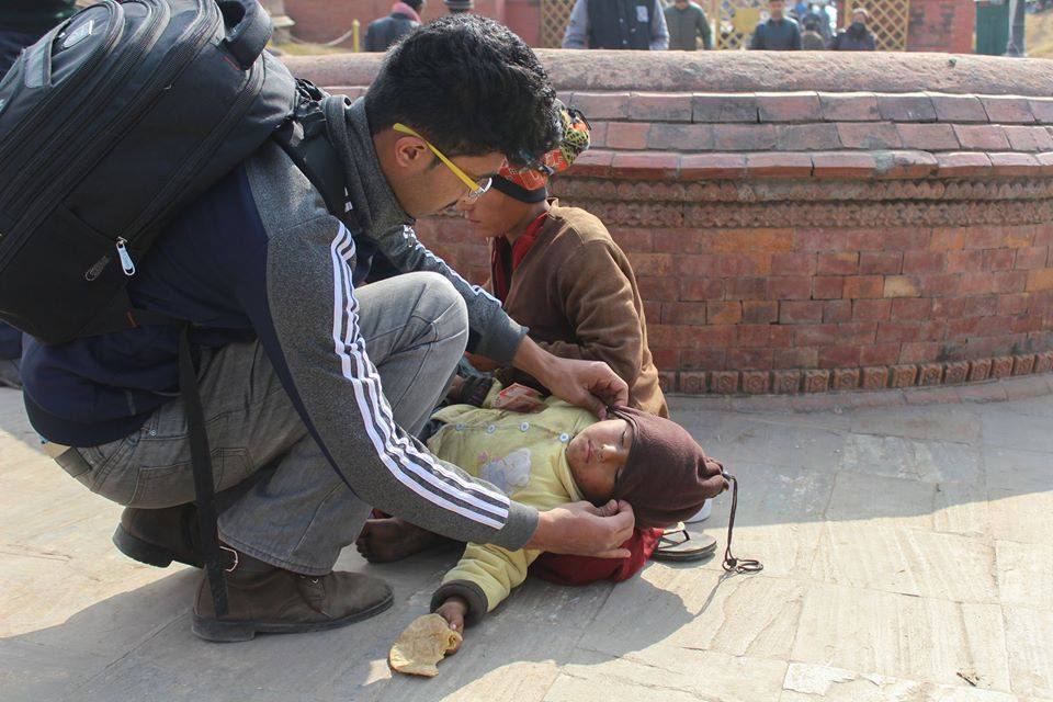 A volunteer kneels on paving to help a man lying on the ground, a temple wall behind them.