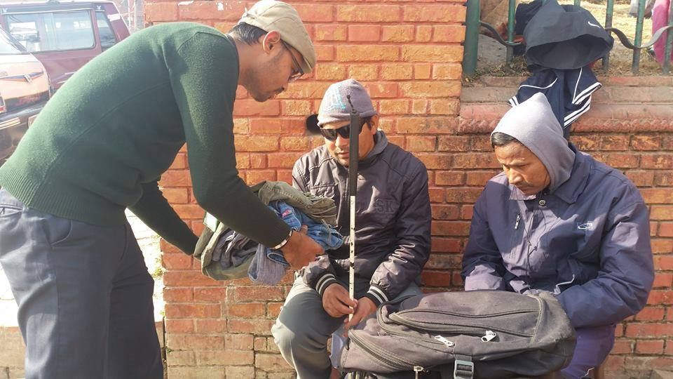 A volunteer in a green jumper bends to hand clothing to two men sitting against a brick wall with a rucksack beside them.