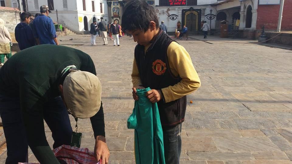 A volunteer bends to give a folded green cloth to a boy in a maroon jumper on a paved street.