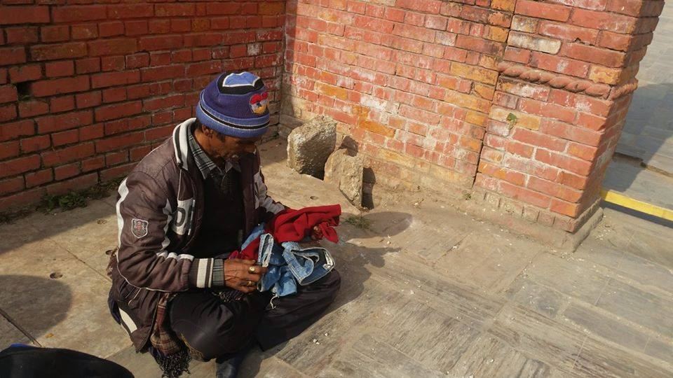 A man crouches on paving beside a brick wall sorting through a pile of clothes, a dog lying nearby.
