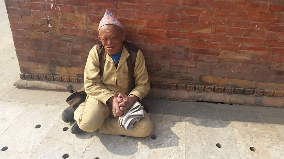 An elderly man in a Dhaka topi sits cross legged against a brick wall on a paved street, holding a bundle in his lap.